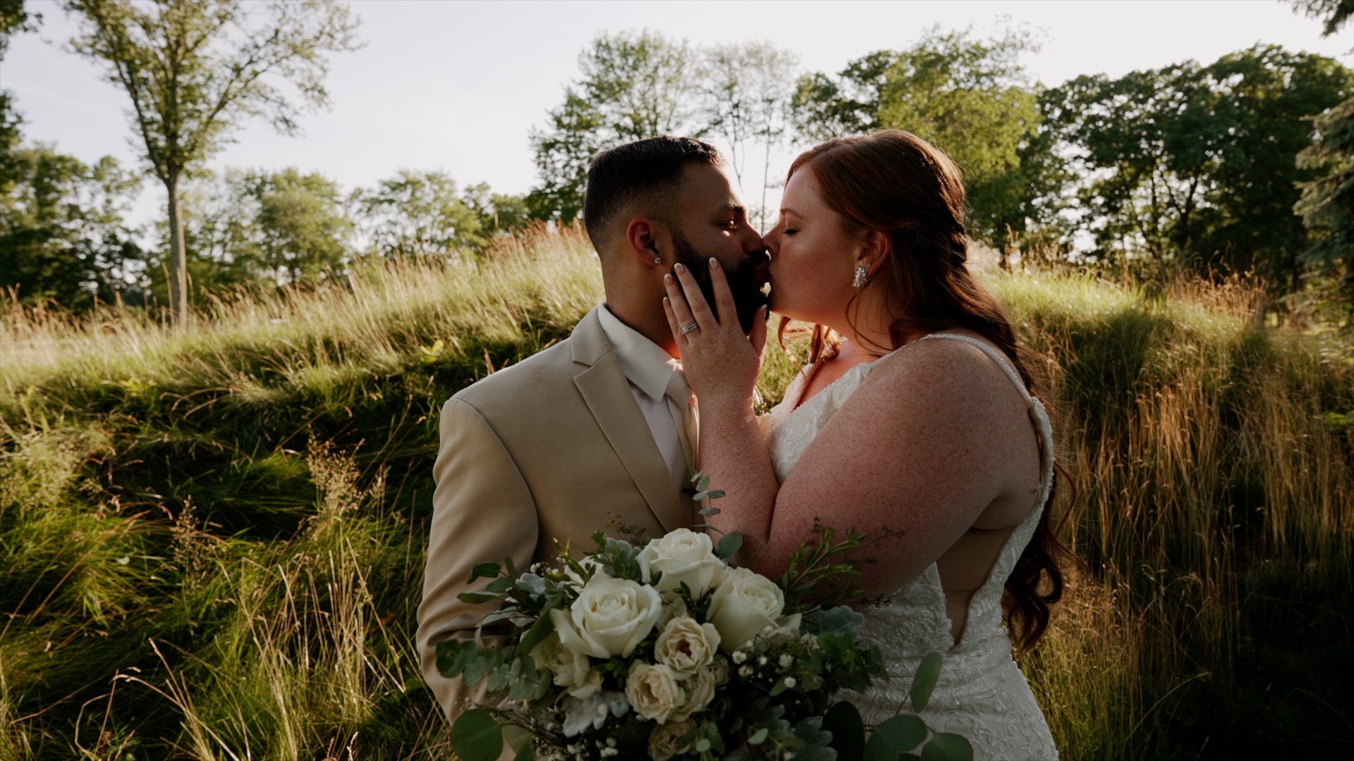 Couple kissing with a hill with tall grass and tree scattered in the background. The bride's left hand is touching the groom's right cheek. You can see the bride's wedding band and engagement ring. Groom is in a tan suit.