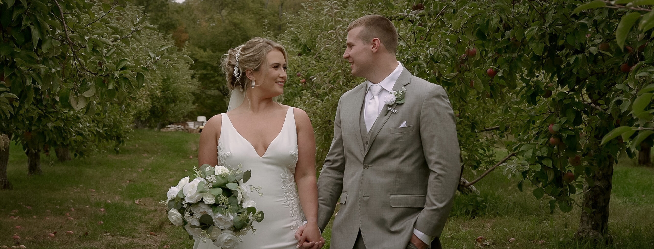 Couple standing side- by- side. holding hands in an apple orchard. Bride is wearing a low cut white dress with lace accents. She has a green and whire bouquet in her right hand and has a cathedral length veil. Groom is wearing a grey suit with a white tie and is standing with his left hand in his pocket