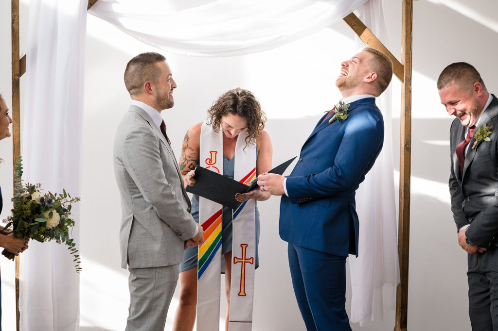 Grooms laughing with their officiant under an arbor at their wedding ceremony