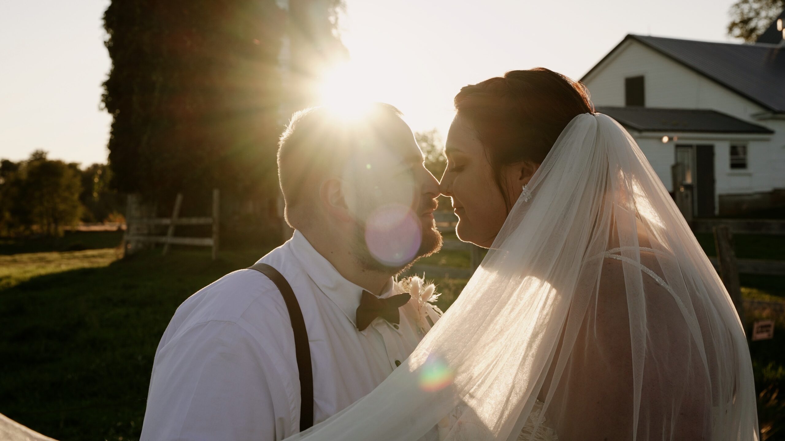 Couple standing in front of a barn and a silo covered with ivy. The sun is setting and causing a solar flare. Their noses are touching. The bride's veil is sweeping from her hair to the left of the frame. Groom is wearing a white button up, suspenders and a bowtie