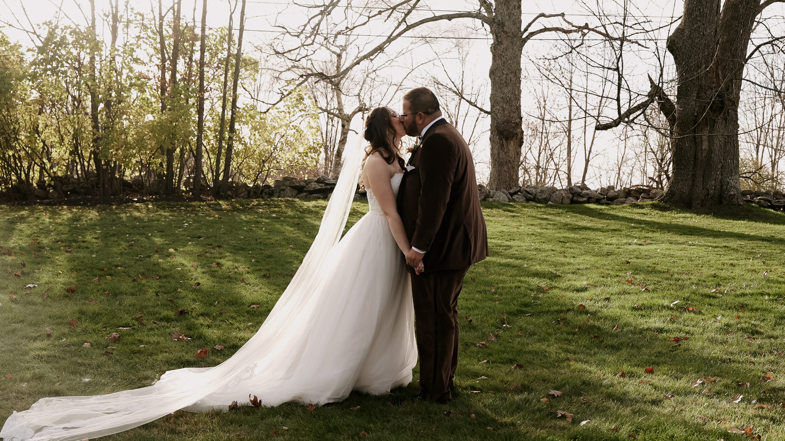 Couple standing in a field kissing. Bride is in a flowy white strapless dress and a long veil. Groom is wearing a brown velvet suit. You can see trees and their shadows and rockwall in the background.