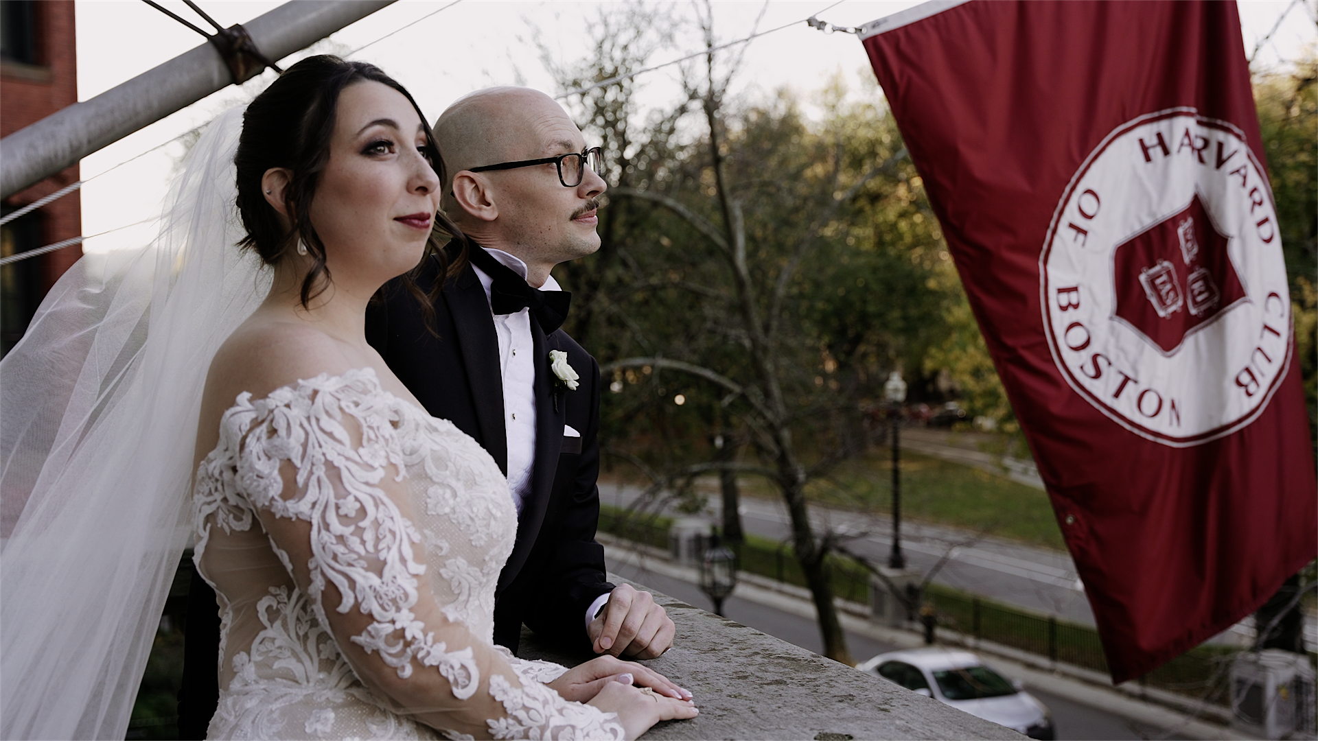 Couple standing on a balcony. The Bride wearing an over-the-shoulder long-sleeved lace dress and a veil and has both hands on the stone railing. Groom is wearing a black tuxedo and is leaning his forearm on the railing. On the right is a maroon-colored flag with a white circle. In the white circle is a maroon insignia and the words "Harvard Club Of Boston" You can see Commonwealth Ave below