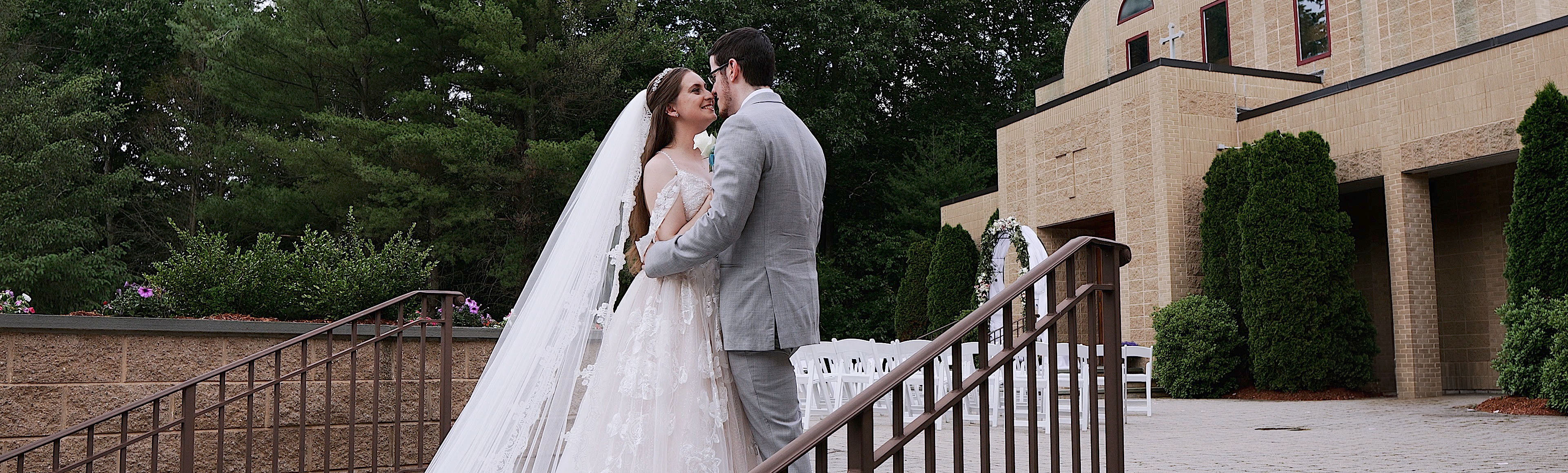 Couple standing on stairs facing each other. You can see a church with an arbor behind the. Groom is wearing a light grey suit and the bride is wearing an almond colored wedding dress with off-the-shoulder straps and has a cathedral length veil.
