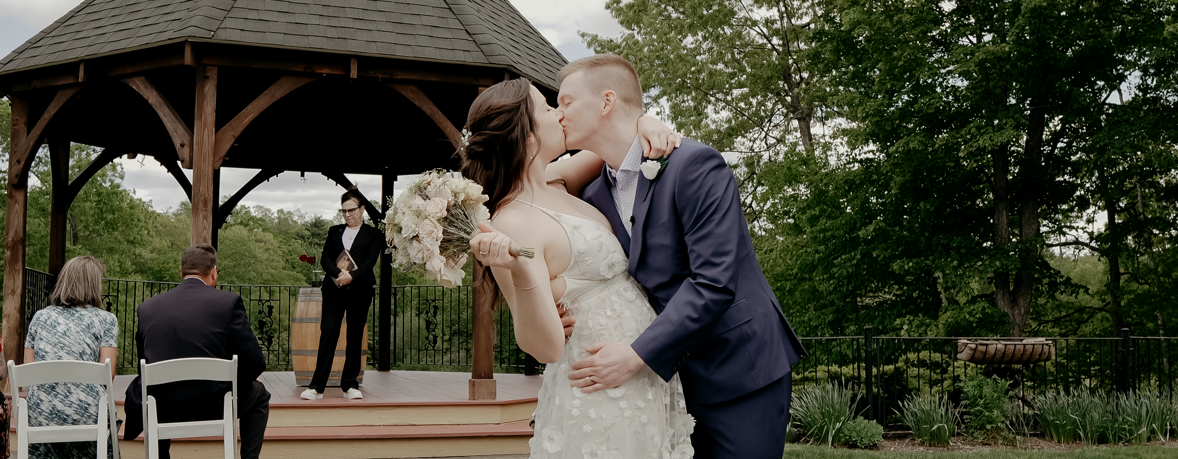 Couple kissing in front of a gazebo. Bride is wearing a white dress with flower accents. She is holding her bouquet up. Groom is wearing a blue suit and has his hands wrapped around her waist.