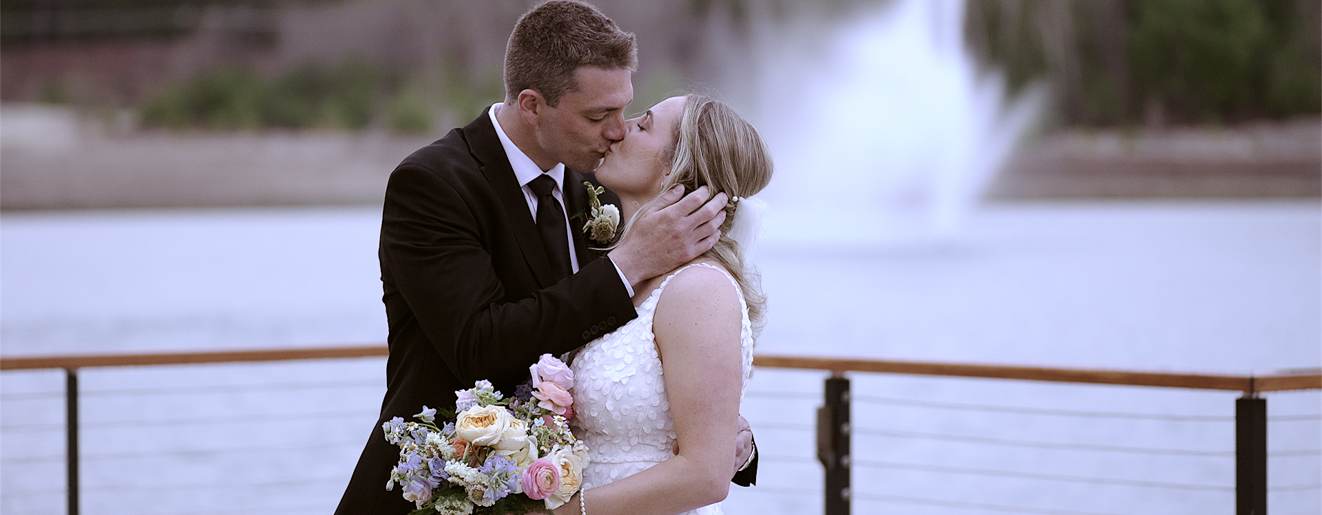 Couple kissing in front of a lake with a fountain. Groom is wearing a black suit and is standing to the right facing the bride. The bride is wearing a white sleevless dress and holding a colorful bouquet. The groom has his right hand up on the left side of the brides face.