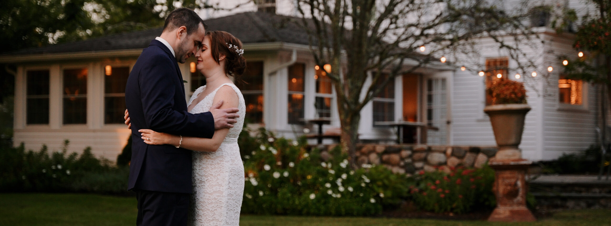 Couple standing facing each other kissing in front of a while house with trees around it. You can see the patio that has bistro lights hanging. Bride is wearing a lace dress that is fitted. Groom is wearing a dark blue suit.