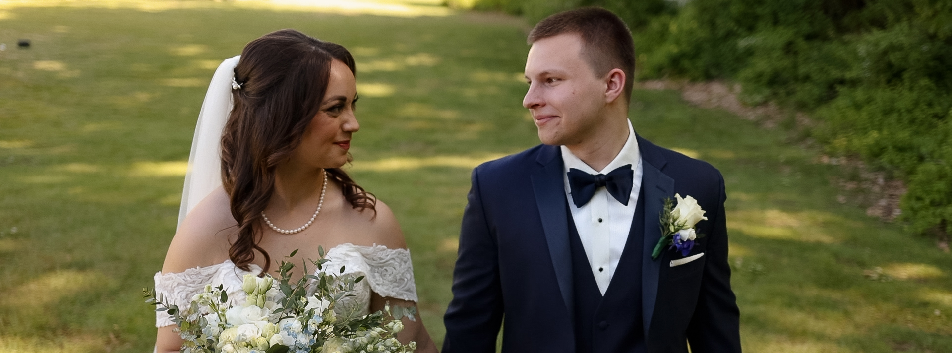 Couple looking at each other while walking in a shady grass field. Bride is wearing a veil, a pearl necklace and an off-the-shoulder dress and the groom is wearing a dark blue suit with a black bowtie