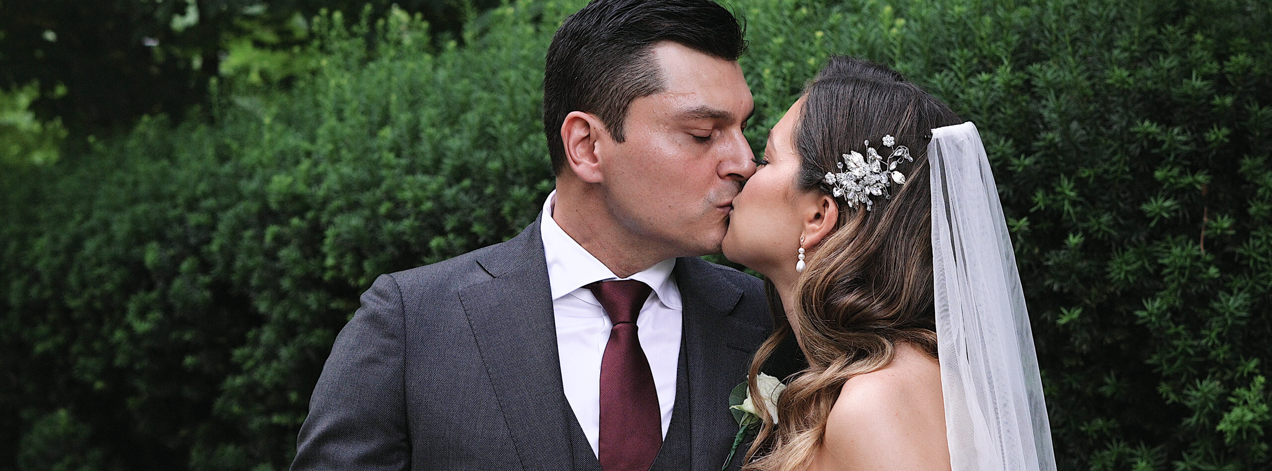Couple standing in front of tall lush bushes kissing with their champagne glasses touching. Groom is wearing a form-fitted dark grey suit with a cranberry colored tie. Bride is wearing a strapless lace dress. She has a diamond covered hair clip over her left ear and is wearing a veil.