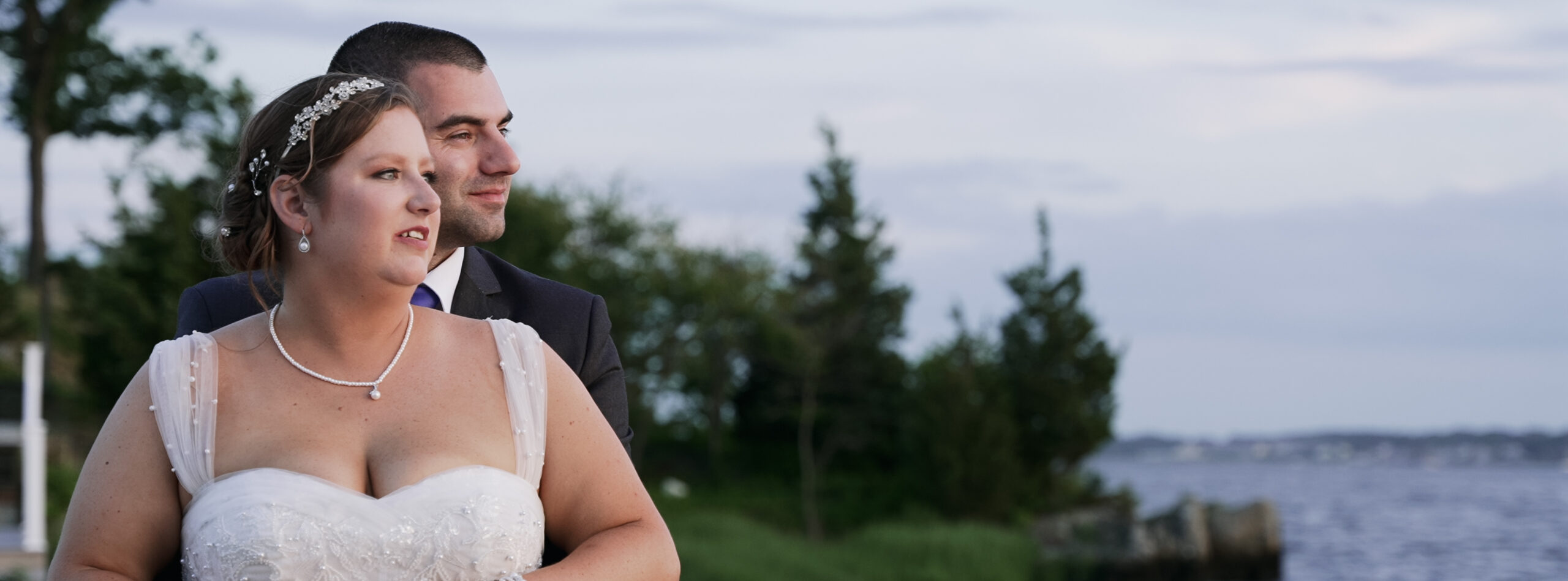 Couple looking of into the distance during their wedding. Groom is wearing a black suit and is standing behind the bride wrapping his arms around her. Bride is wearing a sweetheart dress with straps made out of tule covered in pearls. You can see trees and the bay behind them.