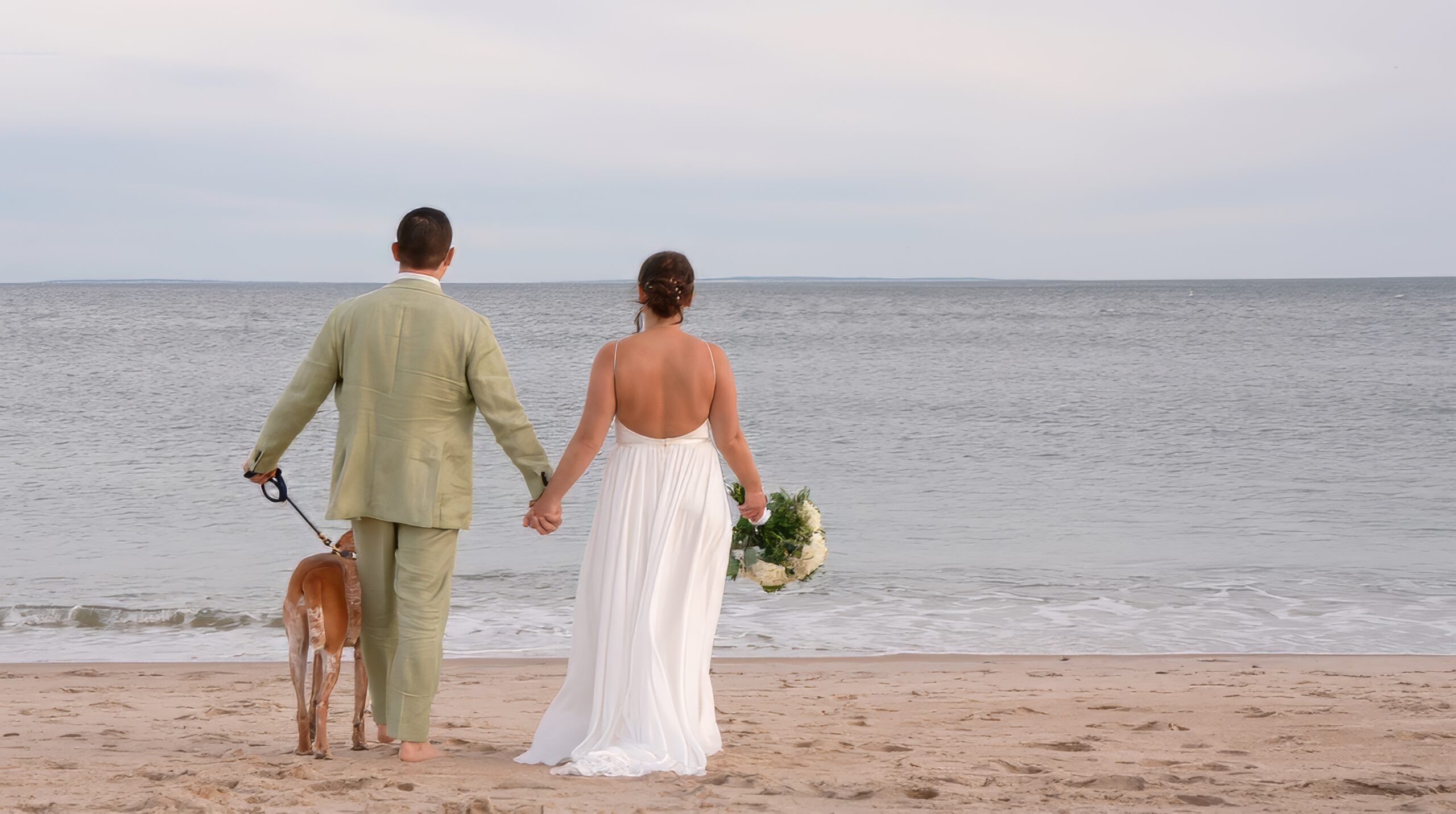 A man in a light green suit walking towards the ocean with a woman in a white dress. To the left the man has a dog on a leash walking beside him. The dog is brown. In the woman's right hand she holds a bouquet to her side
